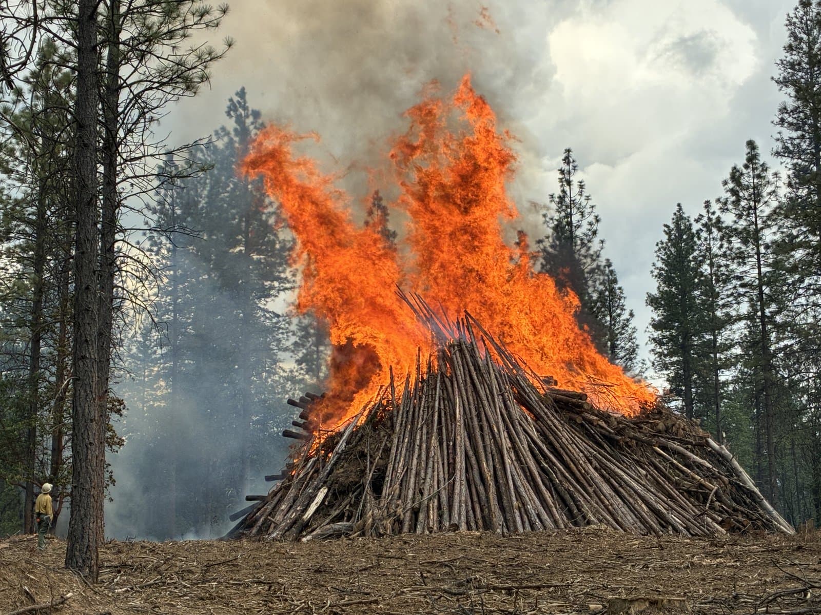 A machine pile burns at full intensity with a firefighter nearby.