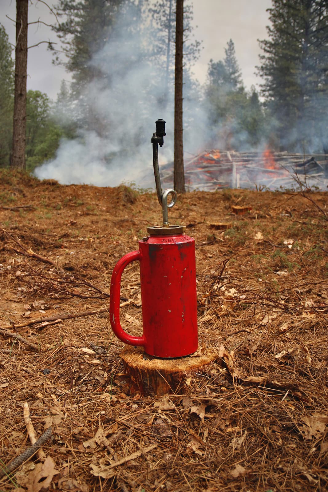 A red drip torch resting on a stump with a smoldering pile and smoky forest behind it. Photo: Hilary Hattie Waters / Yahïla Foto.