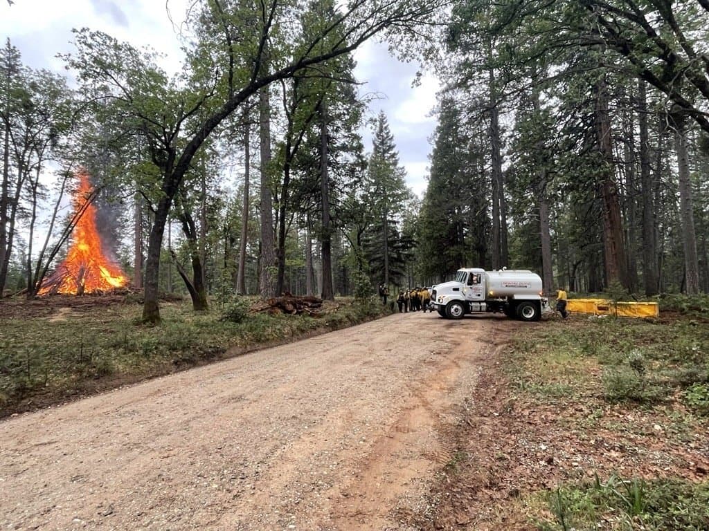 A water tender staged on Jackass Flats Road while a pile burns in an opening beyond the trees.